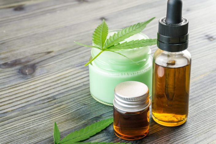 A bottle of CBD oil and a jar of CBD cream are sitting on table. A hemp leaf is on top of the jar.