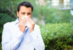 man with allergies outside blowing his nose, allergic to trees in background