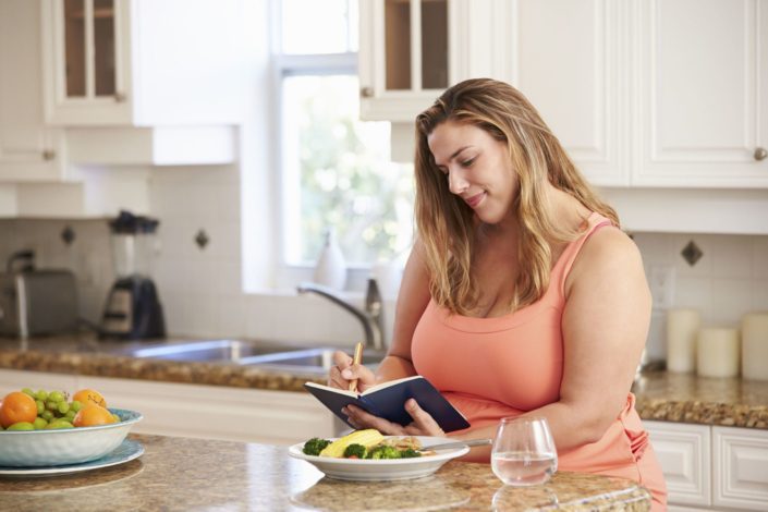 Young, overweight woman eating a healthy meal and writing in a food journal.