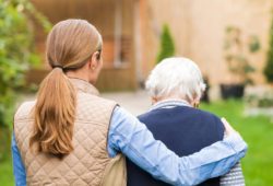 a female caregiver puts her arm around an elderly woman as they walk outside