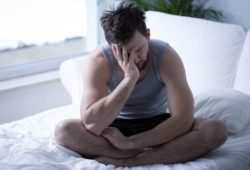 A man who doesn't feel well is sitting on top of his bed with his head in his hand