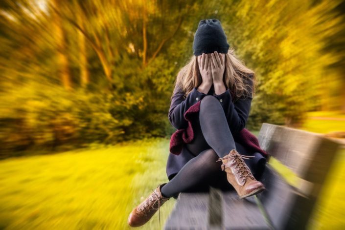 woman sitting on bench outside covering face with hands, surrounded by trees filled with anxiety