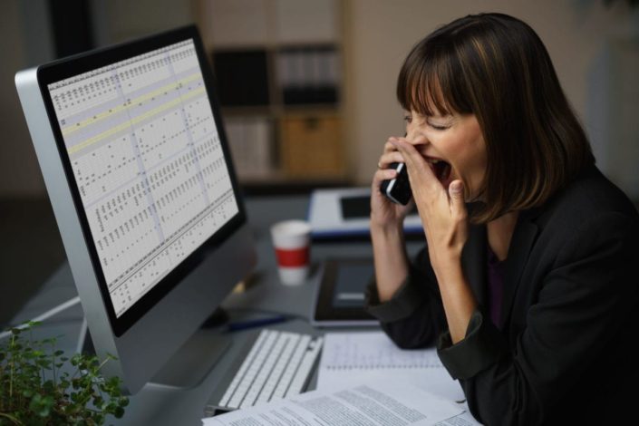 A woman sitting in front of her computer, talking on her cell phone, and yawning