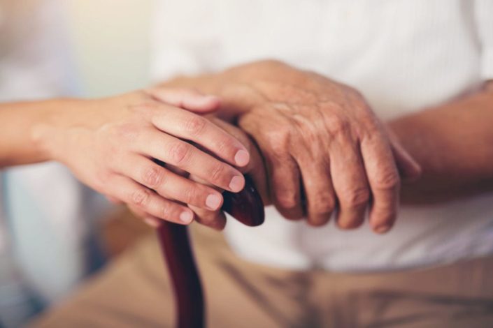 close-up of an elderly man’s hands on top of a cane with a caregiver’s hand on top