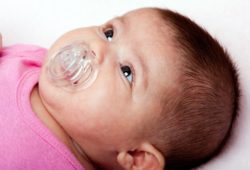 An infant girl sucks on a plastic pacifier in her mouth. Pacifiers have pros and cons, depending upon how long your baby uses one. Your doctor can help you make that decision.