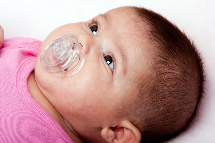 An infant girl sucks on a plastic pacifier in her mouth. Pacifiers have pros and cons, depending upon how long your baby uses one. Your doctor can help you make that decision.