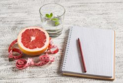 Open food diary next to a grapefruit, glass of water, and measuring tape