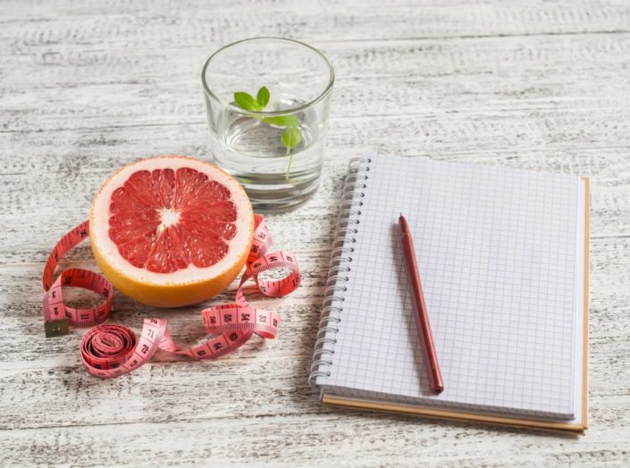 Open food diary next to a grapefruit, glass of water, and measuring tape