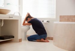 A woman kneeling on the bathroom floor while holding her head over the toilet as she vomits